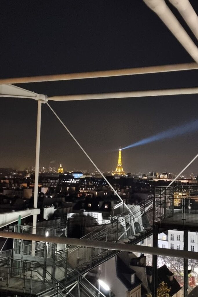 Alt text: Nighttime view from a rooftop bar, showcasing the illuminated Eiffel Tower and the Paris skyline framed by industrial beams and structures. Best rooftop bar in Paris.