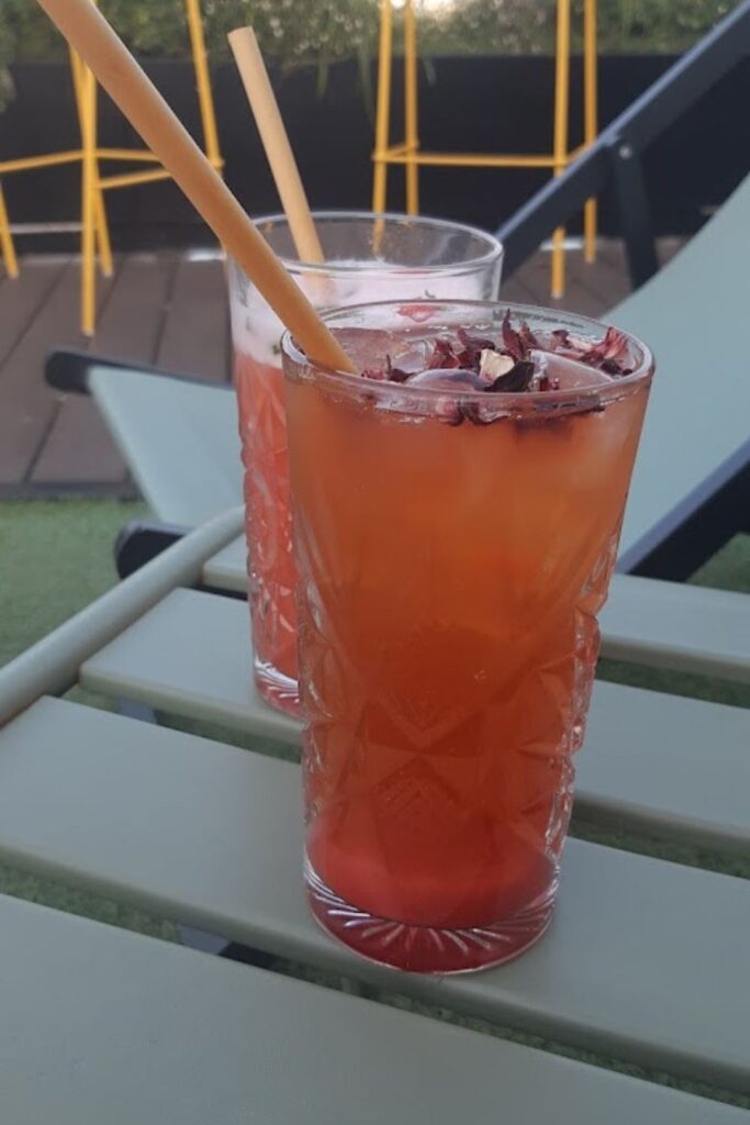 Alt text: Two vibrant cocktails with bamboo straws are placed on a table at a rooftop bar with yellow chairs and greenery in the background. Best rooftop bar in Paris.