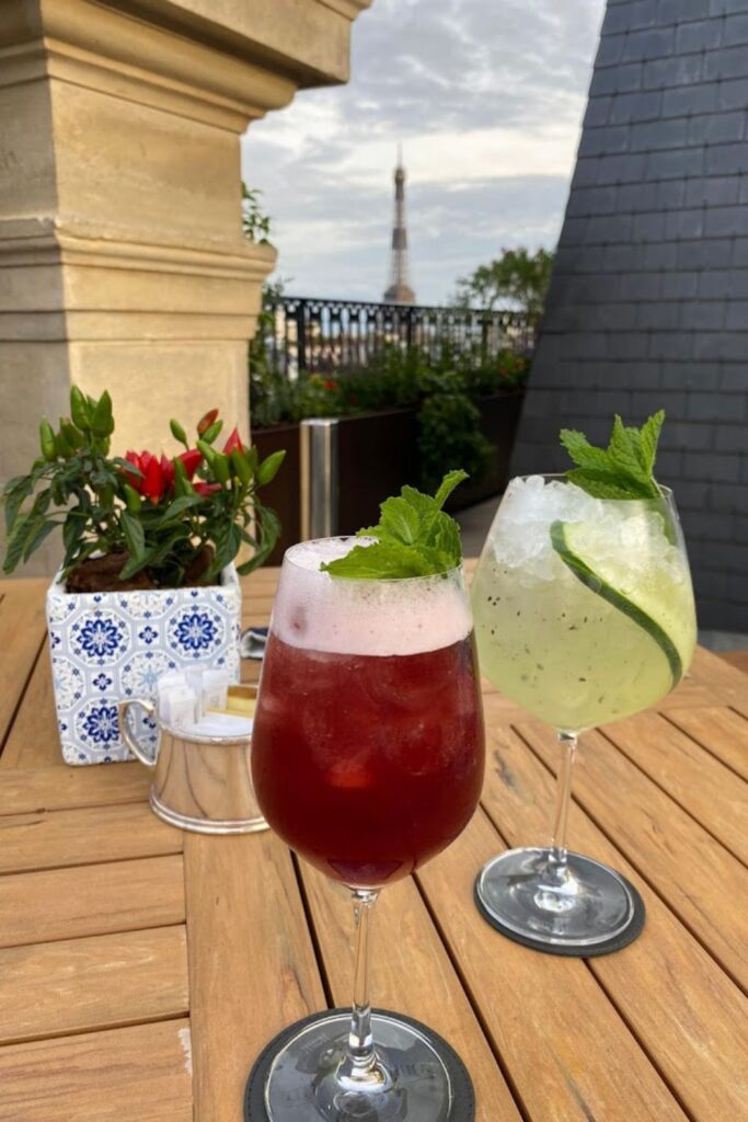 Alt text: Two cocktails, one red and one green, garnished with mint leaves, are placed on a wooden table at a rooftop bar with a view of the Eiffel Tower in the background. Best rooftop bar in Paris.