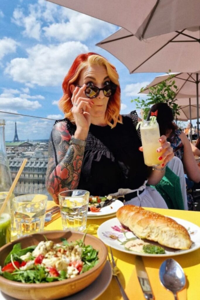 Alt text: A woman with tattoos and sunglasses enjoys a drink at a rooftop bar with food on the table, including a salad and bread, with a view of the Eiffel Tower and Paris skyline in the background. Best rooftop bar in Paris.