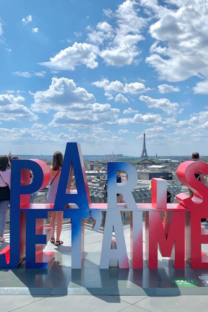 Alt text: People enjoying the view from a rooftop bar with large "Paris Je T'aime" letters in red, white, and blue, overlooking the cityscape of Paris including the Eiffel Tower in the background under a bright blue sky with fluffy clouds. Best rooftop bar in Paris.