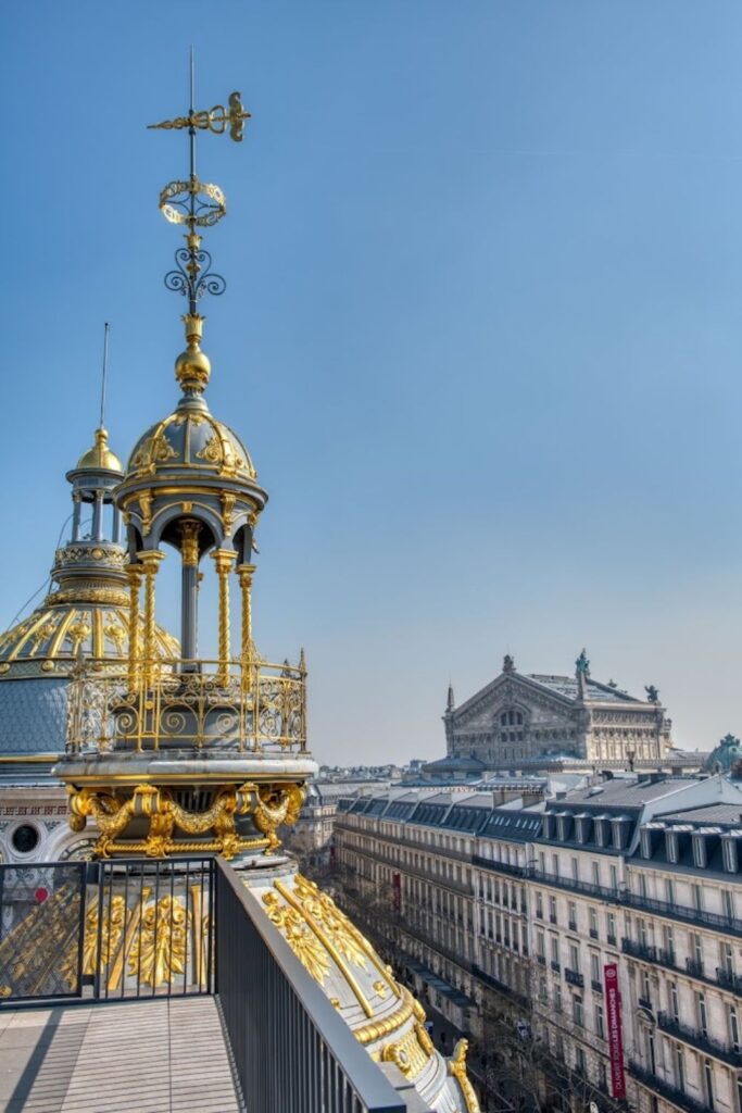 Alt text: View from a rooftop terrace, highlighting a golden, ornate dome and a weather vane against a clear blue sky, with the historic Opera Garnier building in the background. Best rooftop bar in Paris.