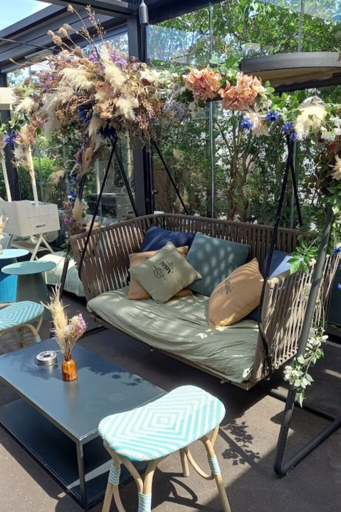 Alt text: A cozy seating area at a rooftop bar with a swing bench decorated with floral arrangements and cushions, surrounded by glass walls and greenery. Best rooftop bar in Paris.