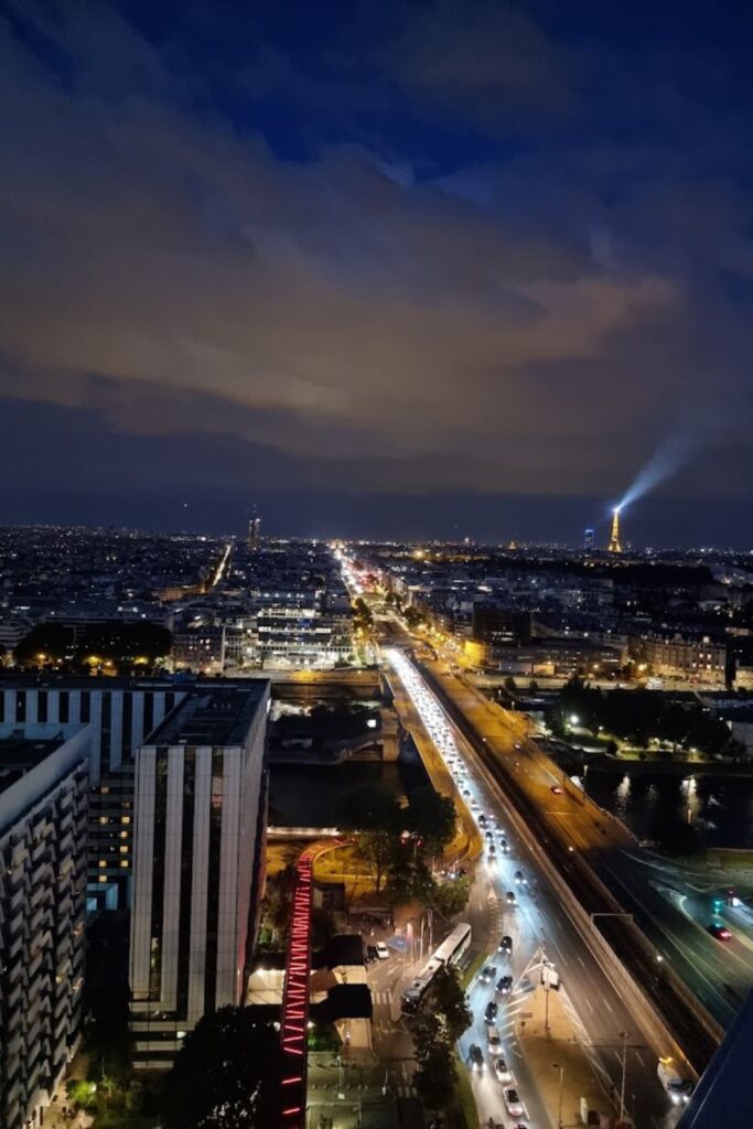 Alt text: Nighttime view from a rooftop bar, showcasing the illuminated Eiffel Tower and a busy city street with car lights streaming into the distance under a partly cloudy sky. Best rooftop bar in Paris.