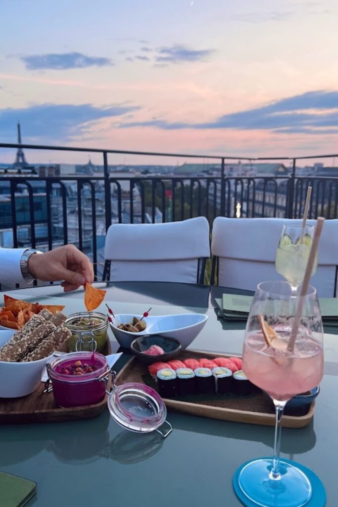 Alt text: A table set with appetizers, including sushi, dips, and breadsticks, and two cocktails at a rooftop bar with a view of the Eiffel Tower and the Paris skyline at sunset. Best rooftop bar in Paris.