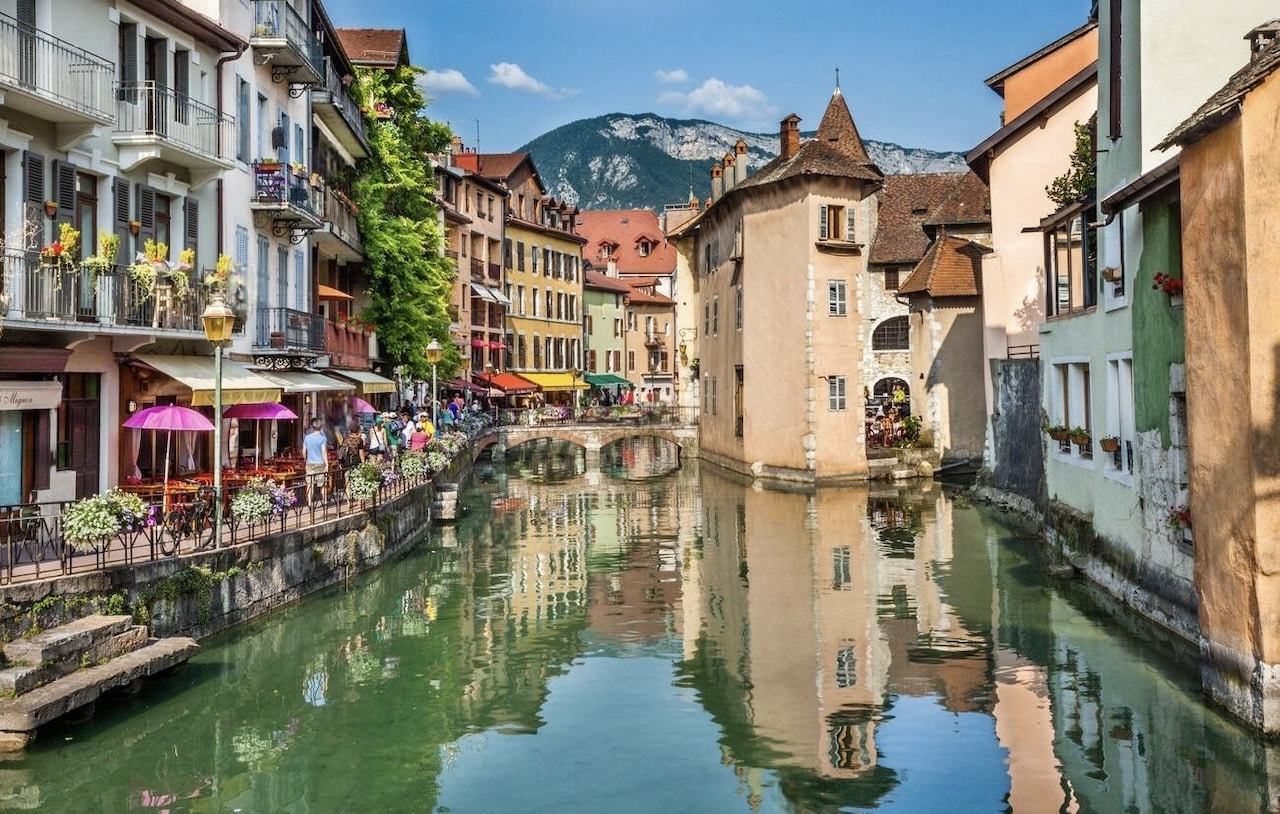 A vibrant scene from one of the prettiest cities in France, Annecy, showcasing the picturesque Thiou Canal. The image captures colorful buildings with flower-draped balconies, charming cafes with outdoor seating, and a quaint stone arch bridge, all set against the backdrop of lush mountains.