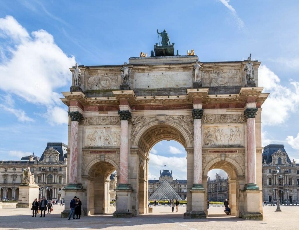 1st arrondissement in paris: Arc de Triomphe du Carrousel in the 1st arrondissement of Paris, showcasing its detailed carvings and statues under a bright blue sky, with the Louvre Museum and the glass pyramid in the background.