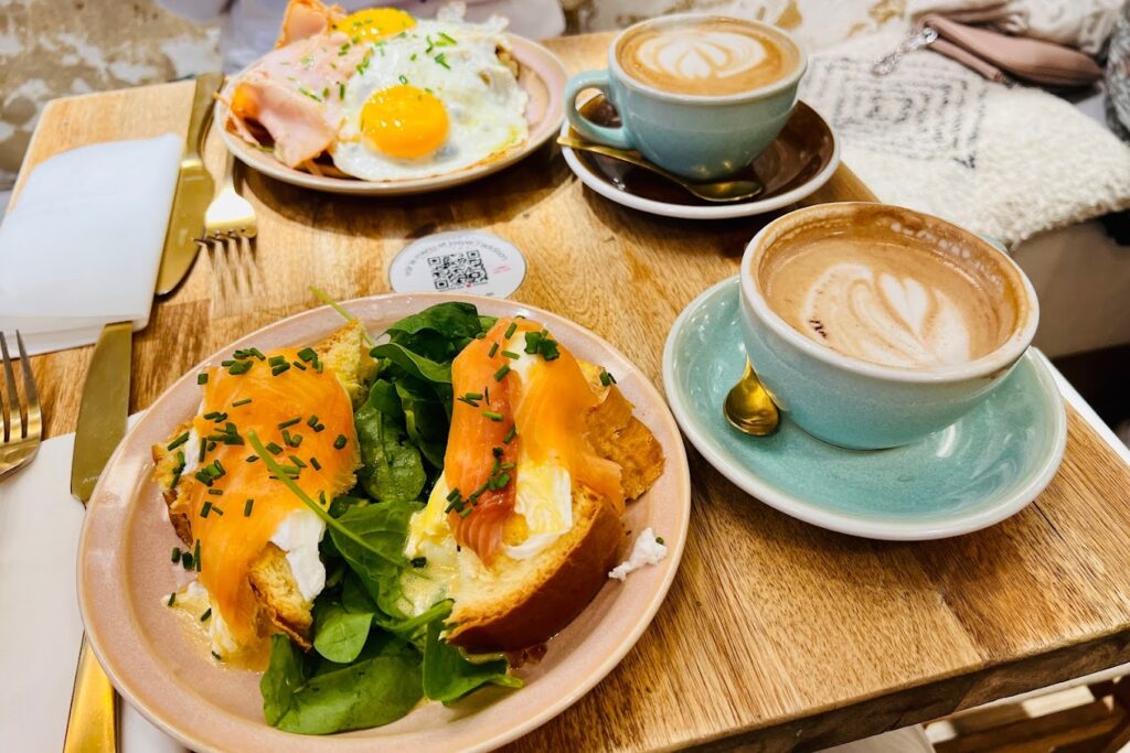 A delectable breakfast spread at Baguett's Café in Paris featuring smoked salmon on toast with cream cheese and fresh greens, alongside a plate of ham and sunny-side-up eggs, complemented by two cups of creamy cappuccino. The setting includes elegant teal and pink crockery on a wooden table, capturing the essence of the best breakfast in Paris.