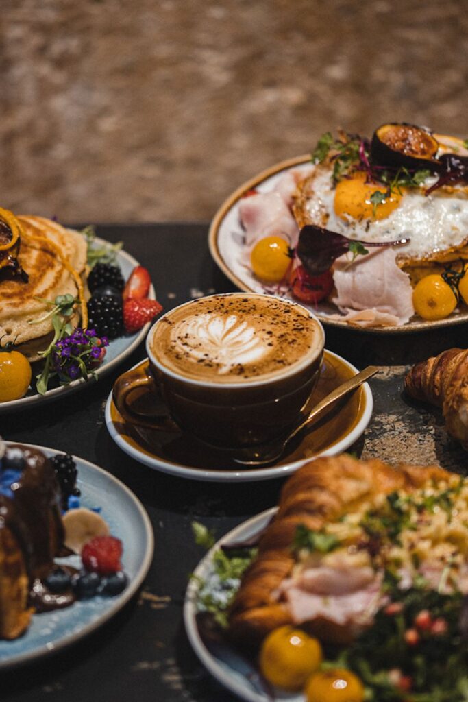 A luxurious breakfast spread at a café near the Louvre in Paris, artfully arranged to capture the essence of the best breakfast in Paris. The meal includes a platter of pancakes garnished with fresh berries and edible flowers, a croissant topped with scrambled eggs and bacon, a French toast adorned with berries and syrup, and a perfectly crafted cappuccino with a delicate foam design. The presentation and variety make it an exemplary Parisian breakfast experience.