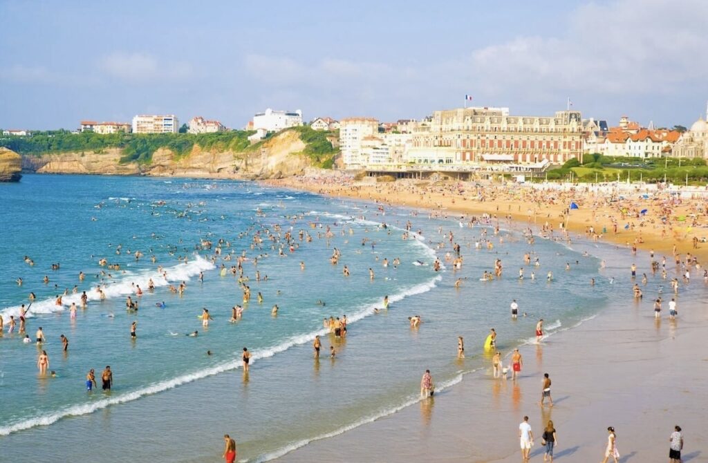 Crowded beach scene in Biarritz, one of the prettiest cities in France, featuring numerous beachgoers enjoying the surf and sand. The coastline is lined with historic and elegant buildings overlooking the vibrant blue sea and golden sandy beach, emphasizing the city's popular and scenic seaside charm.