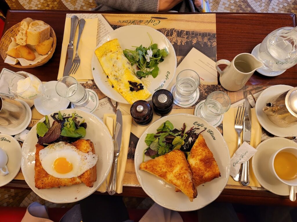 A traditional French breakfast at Café de Flore in Paris, featuring a variety of dishes such as a truffle omelette, croque madame with a sunny-side-up egg, and fresh mixed greens. The table is also set with a basket of fresh baguette slices, butter, jams, and cups of hot tea, arranged meticulously on a table with Café de Flore branded porcelain, capturing the essence of the best breakfast in Paris.