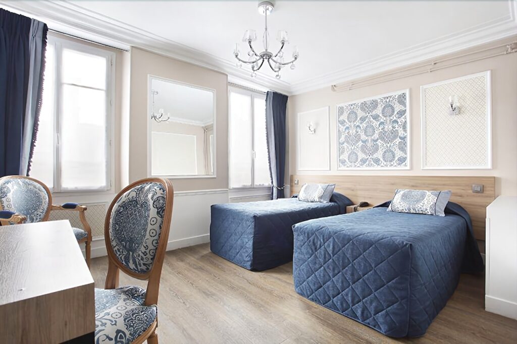 Charming room at Hotel Montpensier, one of the best 1st Arrondissement Paris hotels, featuring twin beds with blue quilted covers, elegant patterned chairs, and classic decor with a chandelier and wall art.