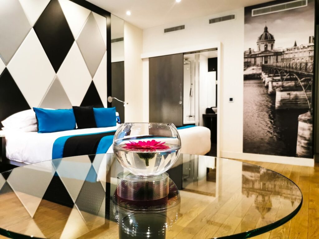 Modern room at L'Empire Paris, one of the best 1st Arrondissement Paris hotels, featuring a stylish bed with blue and black decor, a glass table with a flower centerpiece, and a wall mural of a Parisian bridge.