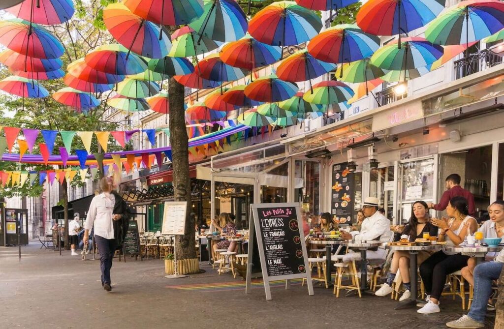 Charming outdoor café scene under a canopy of colorful umbrellas in Le Marais, Paris. Diners enjoy meals at sidewalk tables, surrounded by vibrant decorations and a festive atmosphere, showcasing one of the many delightful things to do in Le Marais.