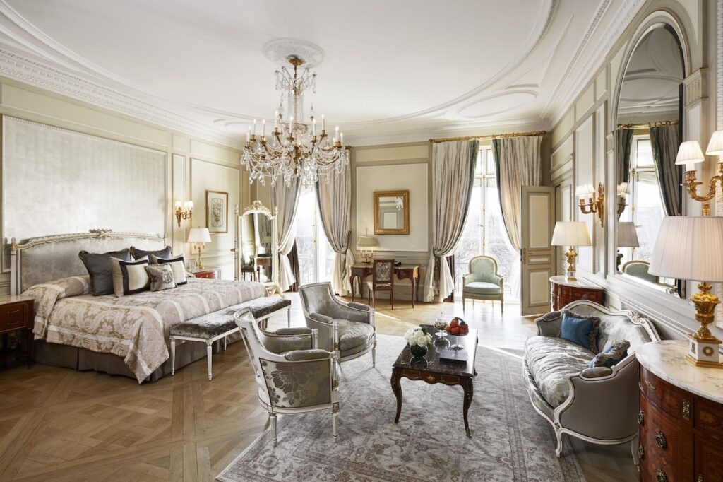 Luxurious room at Le Meurice, renowned as one of the best 1st Arrondissement Paris hotels, featuring opulent decor with a grand chandelier, elegant furniture, and plush bedding in a spacious, light-filled setting.