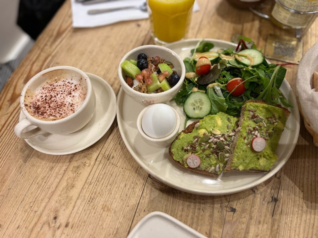 A close-up view of a nutritious breakfast at Le Pain Quotidien in the Marais, Paris, featuring a slice of multigrain toast topped with vibrant green avocado, radishes, and sunflower seeds, alongside a small bowl of mixed fruit salad and a fresh leafy green salad with cherry tomatoes and cucumber. A cappuccino with a sprinkle of cocoa and a glass of orange juice complete this delightful meal, showcasing one of the best breakfasts in Paris.