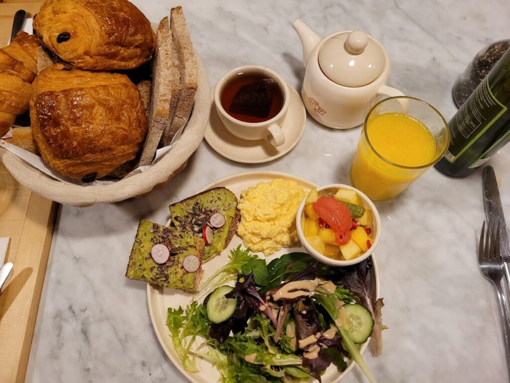 A sumptuous spread from Le Pain Quotidien in the Marais, Paris, showcasing a variety of breakfast items including a basket filled with flaky croissants and assorted breads, avocado toast garnished with radishes, creamy scrambled eggs, a fresh fruit salad, and a green salad. The meal is complemented by a pot of hot tea and a glass of orange juice, capturing the essence of the best breakfast in Paris on a marble tabletop.