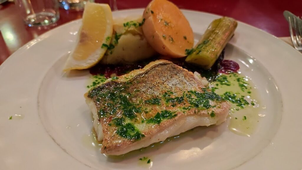 A beautifully plated dish from one of the best restaurants in Bordeaux, France, featuring a fillet of grilled fish drizzled with herb-infused oil, accompanied by roasted vegetables and a wedge of lemon for garnish.