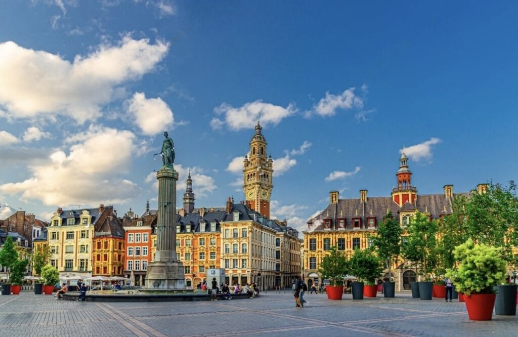 Grand Place in Lille, recognized as one of the prettiest cities in France, with its striking blend of Flemish architecture. The square is dominated by the towering belfry of the Chamber of Commerce, a bronze statue in the foreground, and colorful historic buildings under a vivid blue sky with fluffy clouds.