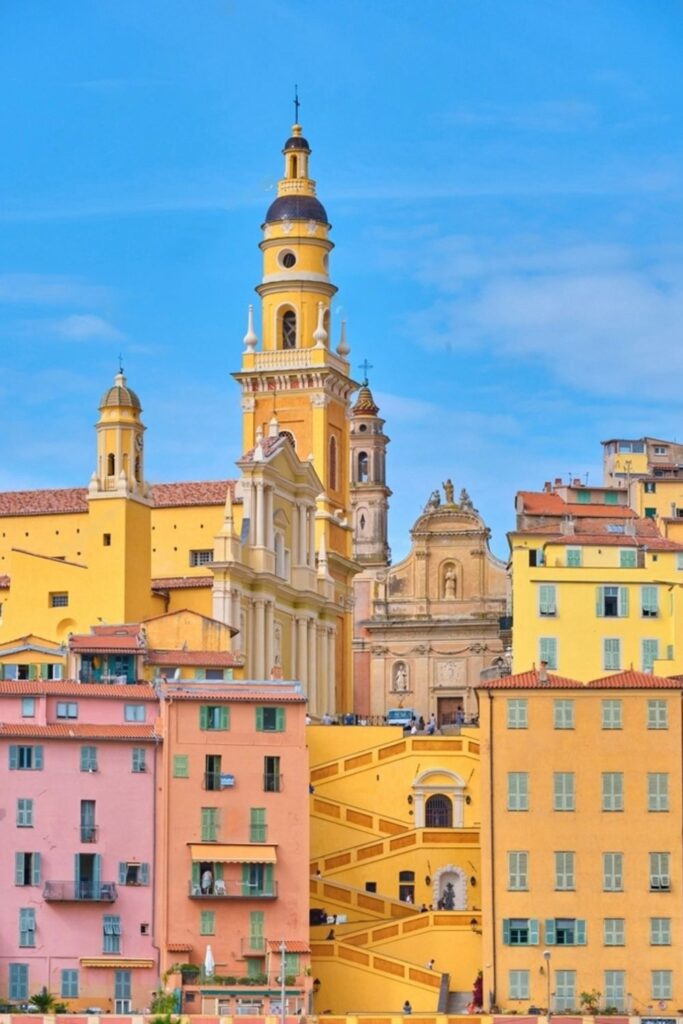 Stunning view of the pastel-colored buildings and intricate architecture of Menton, a top choice for best day trips from Nice, featuring the striking yellow façade and ornate bell tower of the Basilica of Saint Michel.