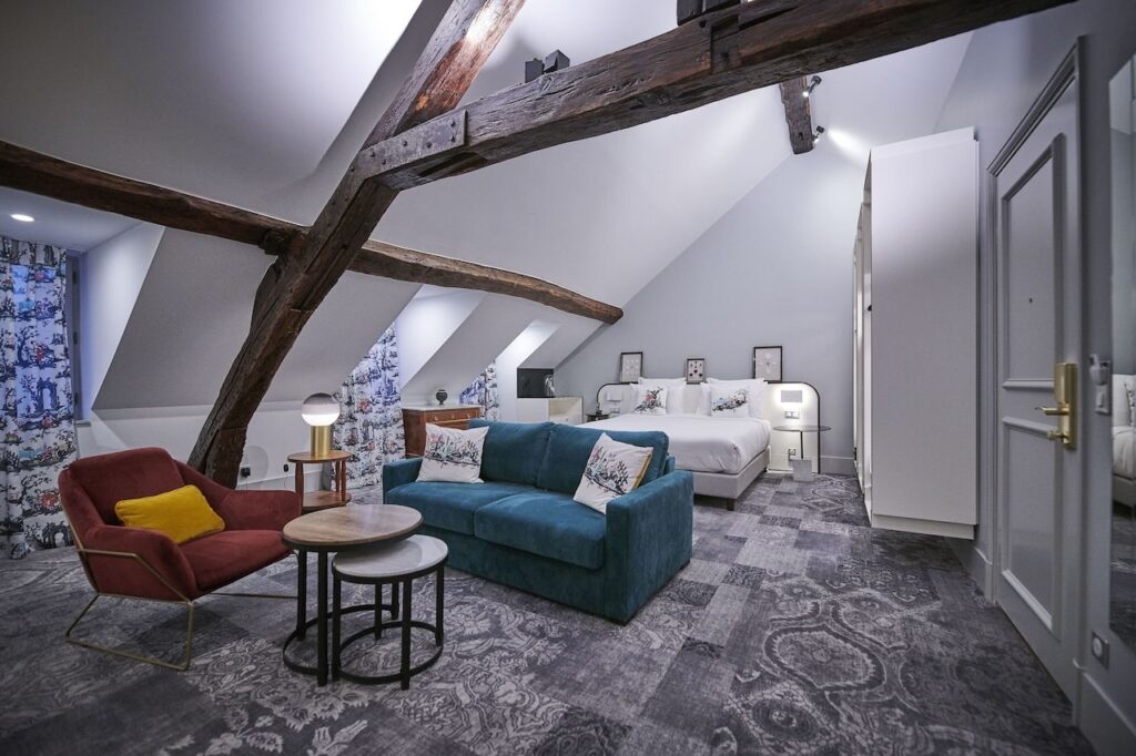 Spacious loft-style room at Normandy Le Chantier, one of the best 1st Arrondissement Paris hotels, featuring exposed wooden beams, a comfortable seating area, and a cozy bed with artistic decor.