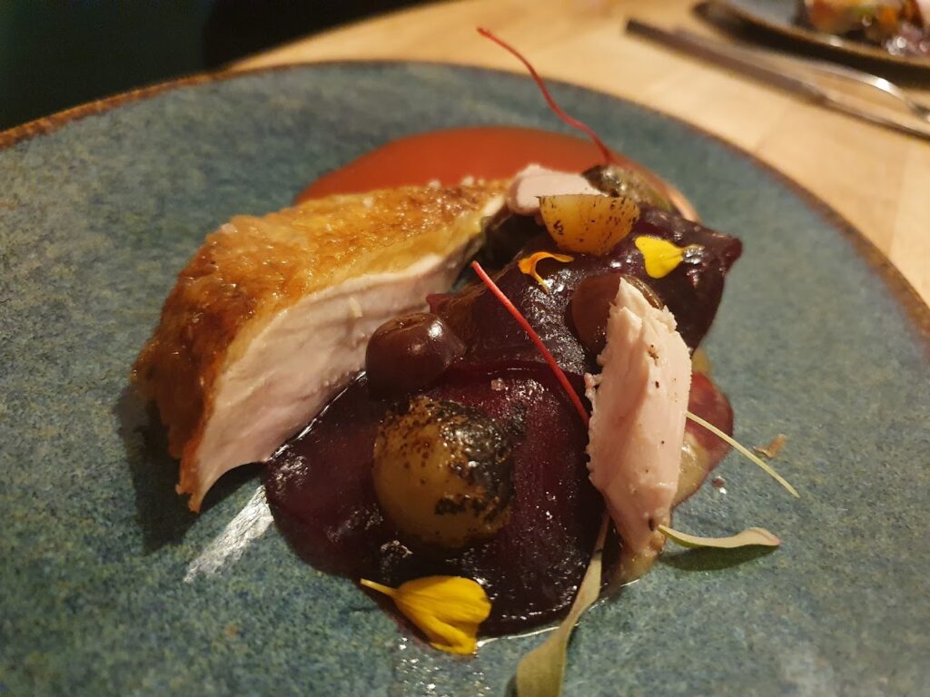 A beautifully crafted dish from one of the best restaurants in Bordeaux, France, featuring a succulent piece of roasted poultry with crispy skin, accompanied by glazed vegetables, vibrant beet puree, and delicate edible flowers.