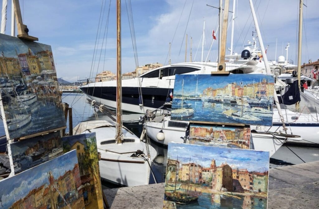 Artistic display of painted canvases depicting the vibrant harbor of Saint-Tropez, a prime location for the best day trips from Nice, with real luxury yachts and sailing boats moored in the background, blending art and reality along the sunlit waterfront.