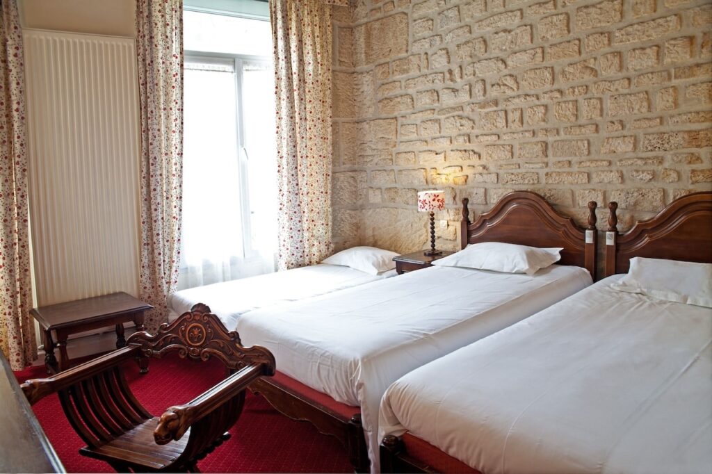 Cozy room at Tonic Hotel Du Louvre, regarded as one of the best 1st Arrondissement Paris hotels, featuring three single beds with classic wooden headboards, floral curtains, and an exposed stone wall.
