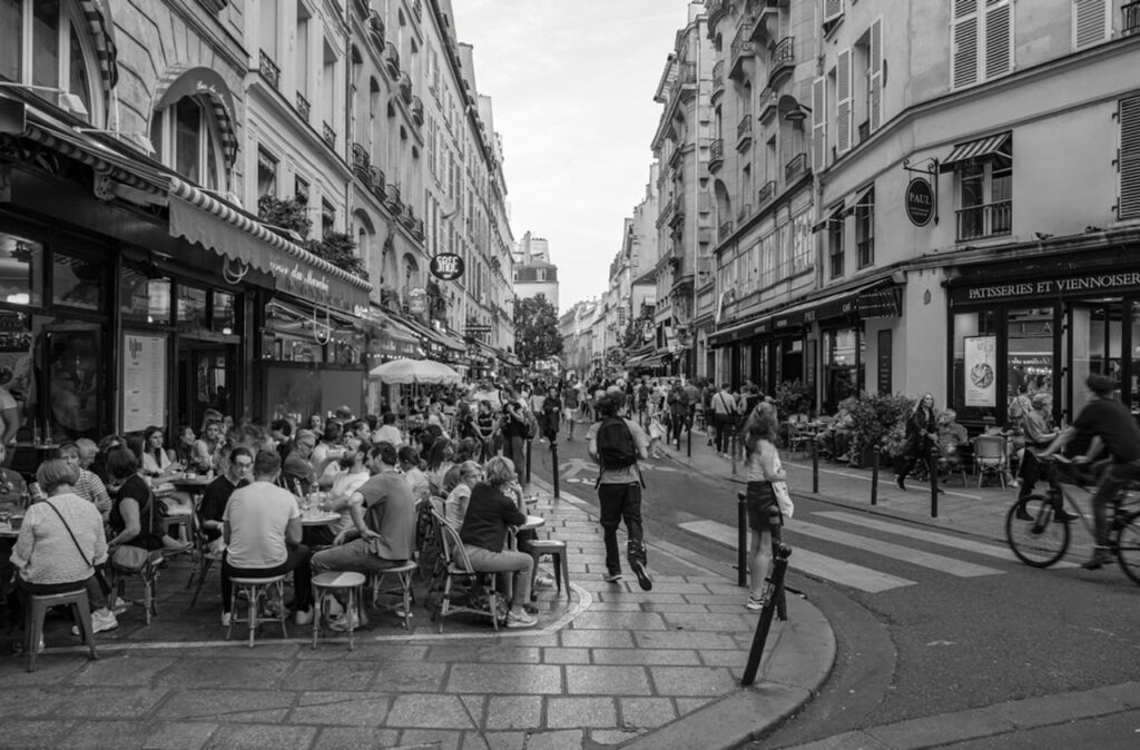 A black and white photograph of a lively street scene in Paris, featuring crowds of people dining at outdoor cafés and walking along a narrow road flanked by historic buildings. Cyclists and pedestrians merge seamlessly in the dynamic environment, embodying the vibrant street culture ideal for Instagram quotes about the spirited Parisian lifestyle.