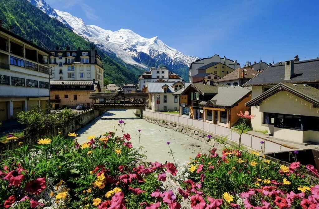 Summer view of Chamonix, nestled among the French Alps and regarded as one of the prettiest cities in France. The foreground is adorned with vibrant flower beds in full bloom, overlooking a tranquil river that flows through the town with traditional alpine buildings and snow-capped mountains in the background.