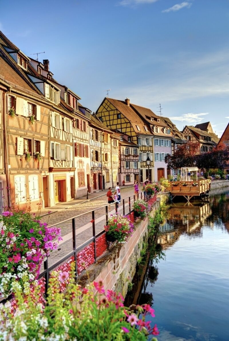 Scenic summer view of Colmar, celebrated as one of the prettiest cities in France, featuring a canal lined with vibrant flowers and traditional half-timbered houses. Pedestrians stroll along the cobblestone pathways, enjoying the charming and colorful architecture reflected in the calm water.