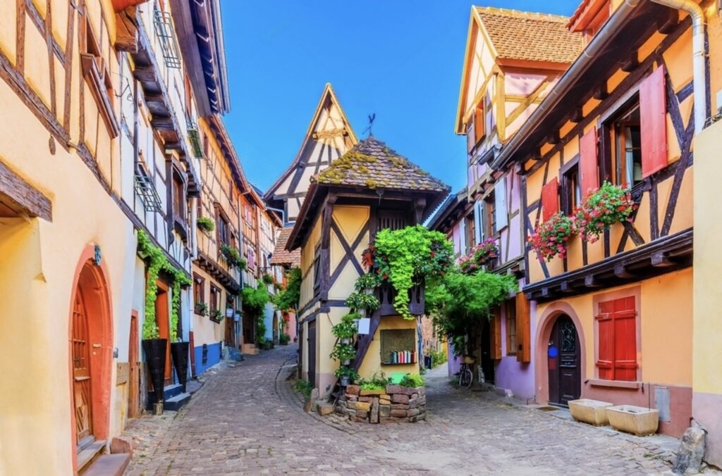Quaint cobblestone street in Eguisheim, one of the prettiest cities in France, lined with colorful half-timbered houses adorned with flower-filled window boxes. A distinctive well house covered in greenery sits at a street corner, enhancing the medieval charm of this picturesque village.