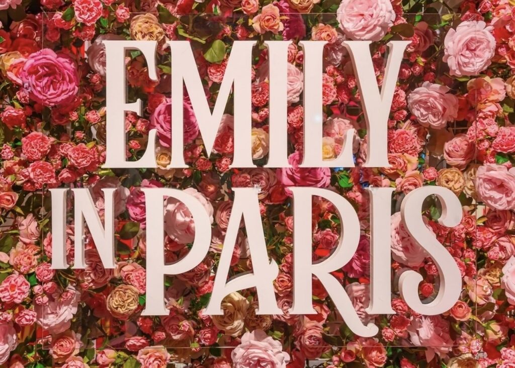 The words 'Emily in Paris' in large, bold, white letters centered on a dense backdrop of vibrant roses in various shades of pink and red. This vivid display captures the romantic essence of Paris, perfect for Instagram quotes celebrating the city's charm and allure.
