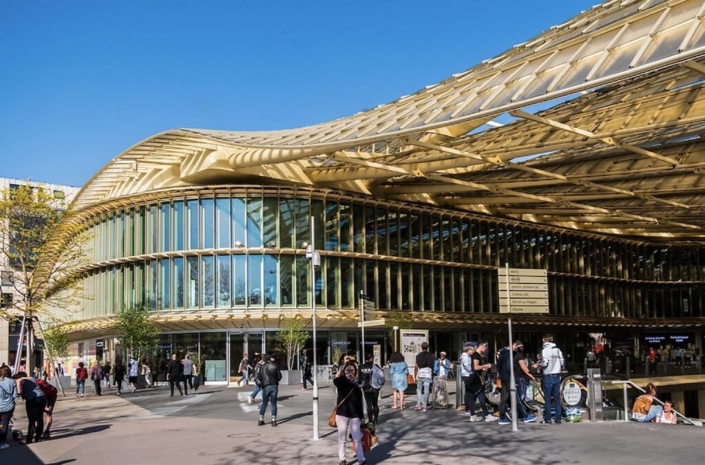 1st arrondissement in paris: the exterior of Forum des Halles in Paris, featuring its modern architectural design with a wavy, gold-colored roof and large glass windows. People are gathered and walking around the plaza in front of the building under a clear blue sky.