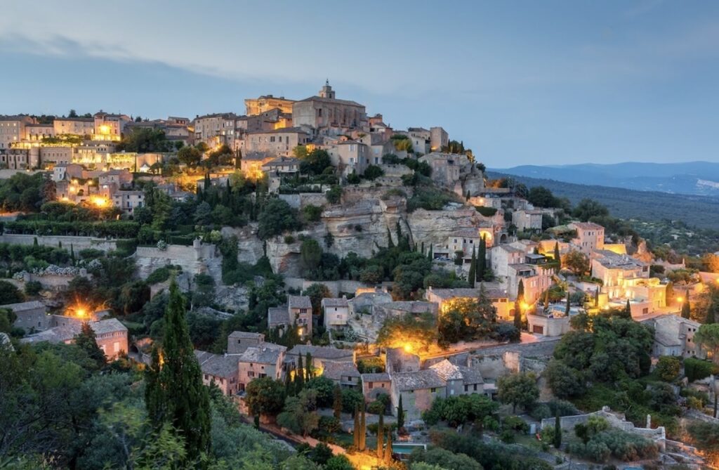 Twilight view of Gordes, a medieval hilltop village renowned as one of the prettiest cities in France. The stone buildings, perched on a steep hill, are illuminated by warm lights that highlight their historic charm against the backdrop of the Provencal landscape and a dusky sky.
