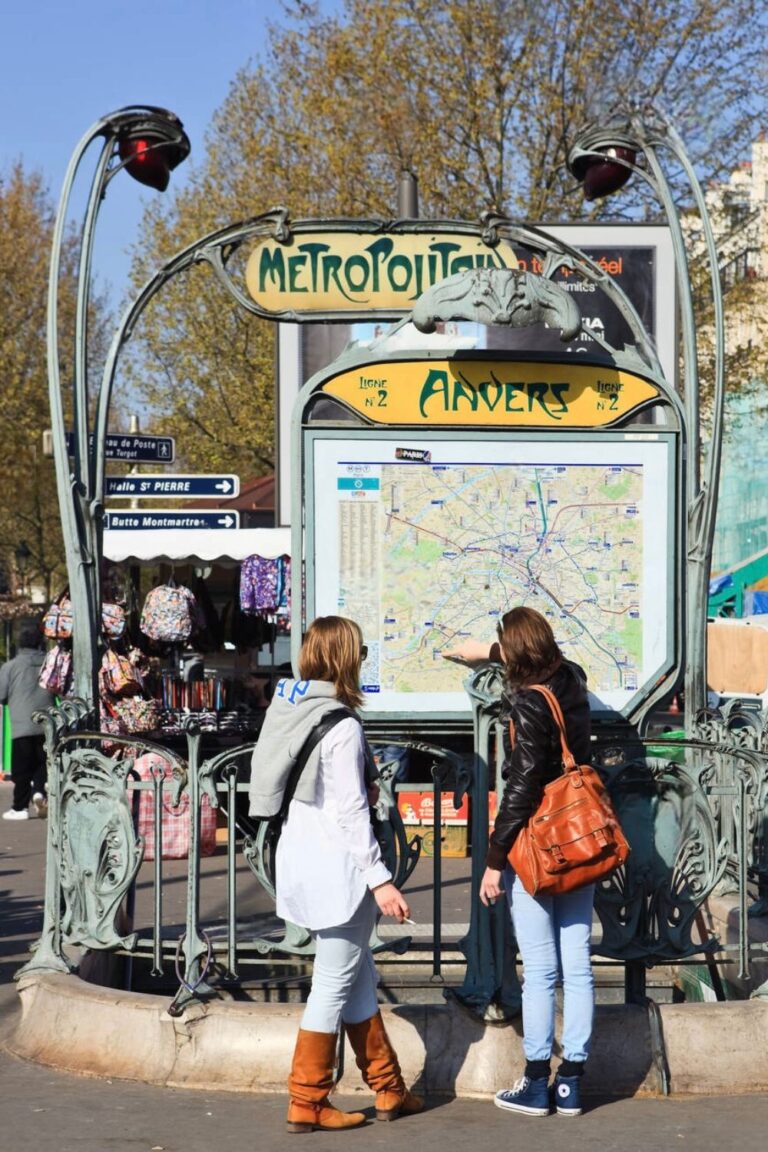 Two people are looking at a map at the Anvers metro station entrance in Paris. The entrance features iconic Art Nouveau design elements, including ornate green metalwork and a sign reading "Metropolitain."