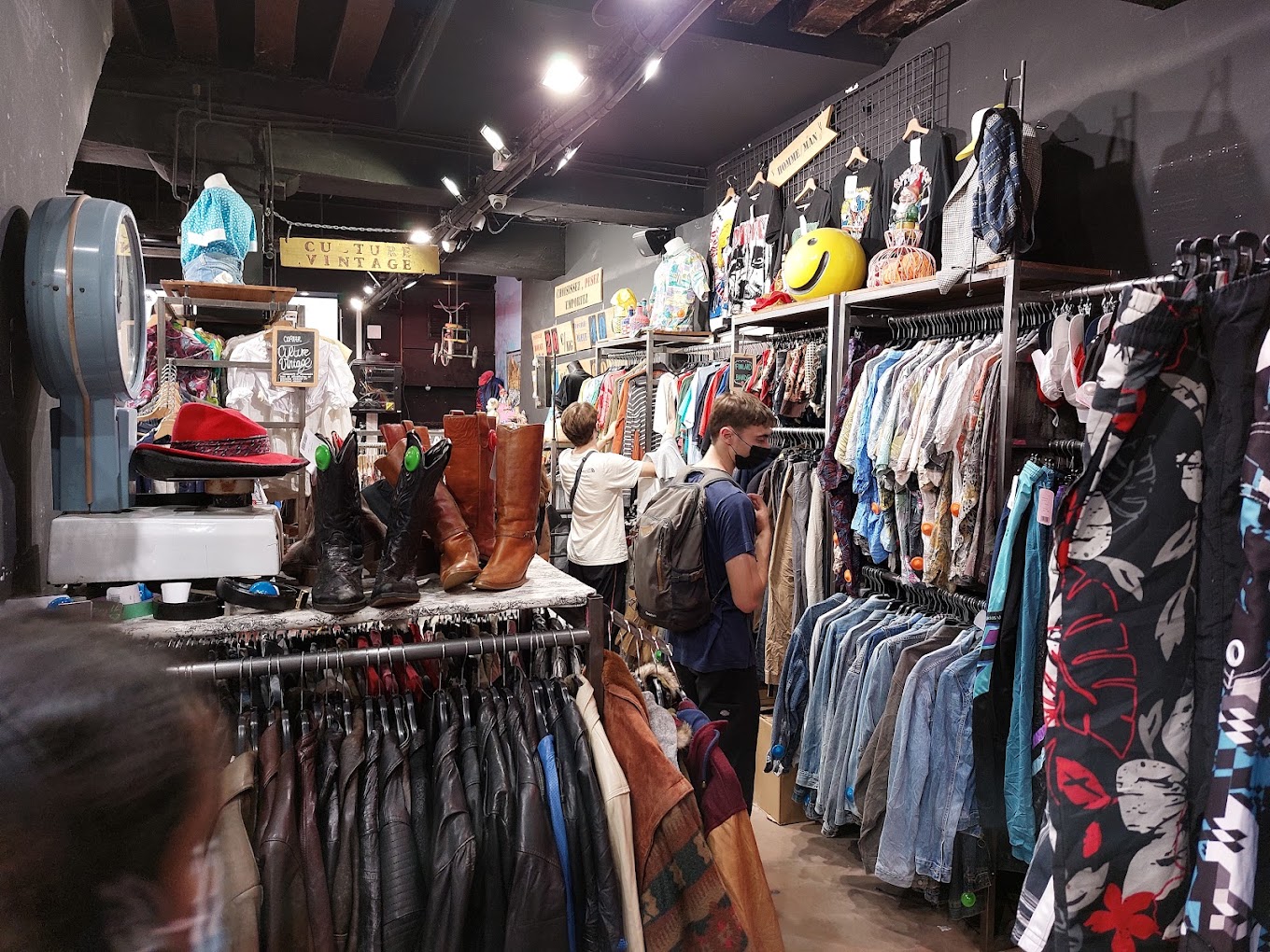 Interior of the Kilo Shop, a popular vintage store in Le Marais, Paris. The scene shows diverse shoppers browsing through an eclectic mix of items, including racks of colorful vintage clothing and shelves filled with boots and accessories, creating a vibrant and bustling shopping atmosphere.