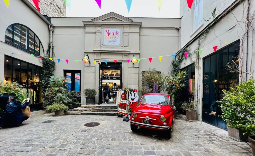 Quaint courtyard view at Merci, a concept store in Le Marais, Paris. The scene features a classic red Fiat car parked in front of the store, surrounded by festive bunting and a welcoming, artistic atmosphere, offering a glimpse into the unique shopping things to do in Le Marais.
