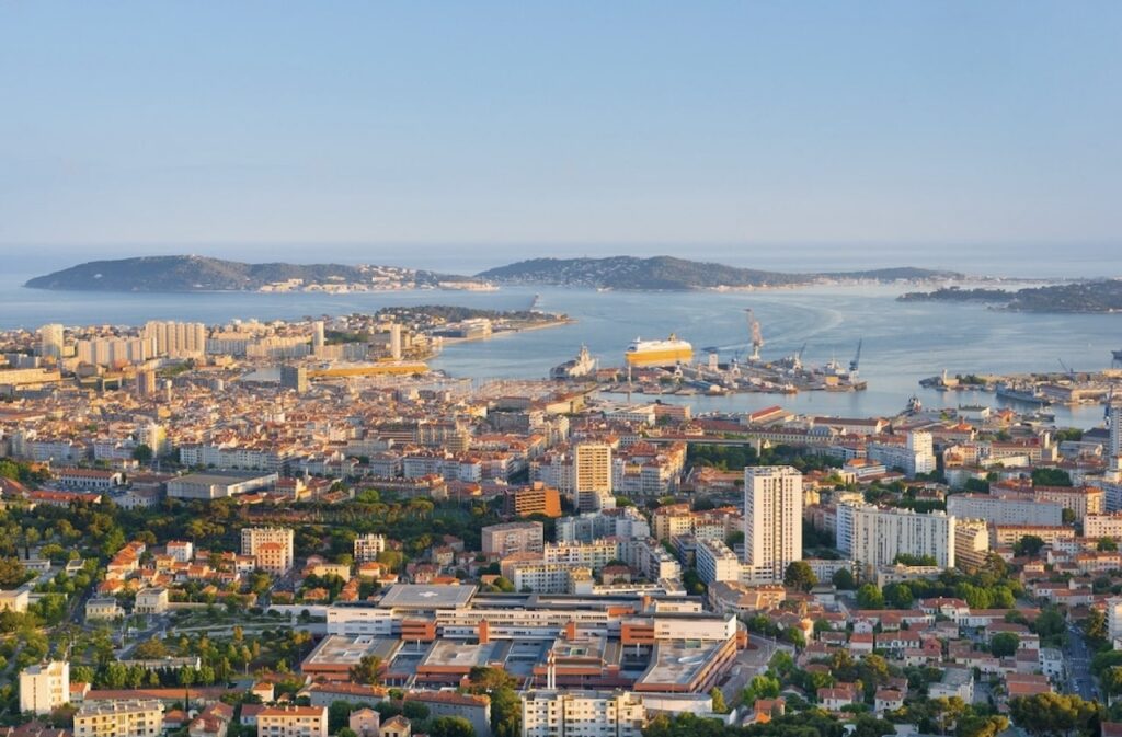 Panoramic view of Toulon, France, a popular choice for the best day trips from Nice, featuring densely packed urban architecture with a mix of modern and traditional buildings, and large passenger ships docked in a bustling harbor.