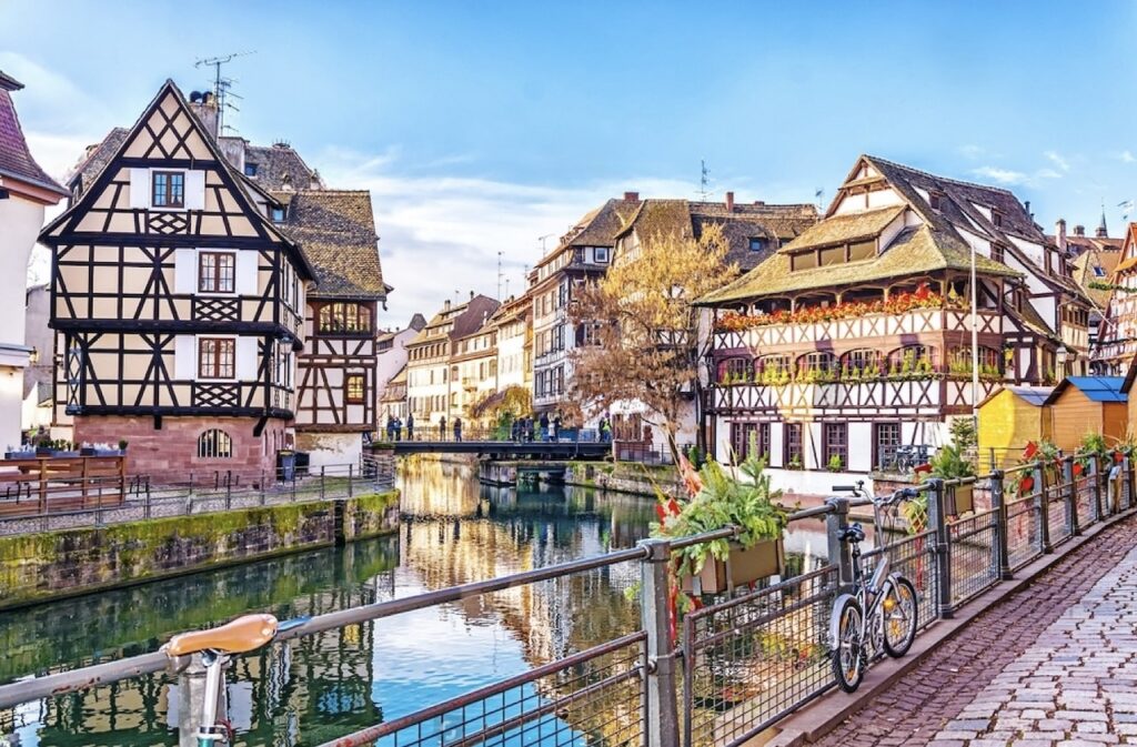 Picturesque canal view in Petite France, Strasbourg, one of the prettiest cities in France. The scene features charming half-timbered houses with colorful shutters and flower boxes, a calm canal reflecting the vibrant buildings, and a bicycle parked along the cobbled street, enhancing the old-world ambiance of the district.
