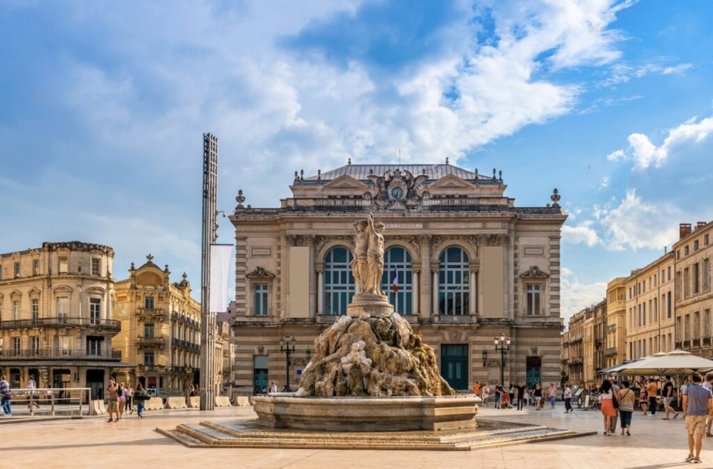 The Place de la Comédie in Montpellier, one of the prettiest cities in France, bustling with activity under a bright blue sky. The square features the elegant Opera House with classical architecture and a striking fountain statue at its center, surrounded by historic, ornate buildings and lively street scenes.