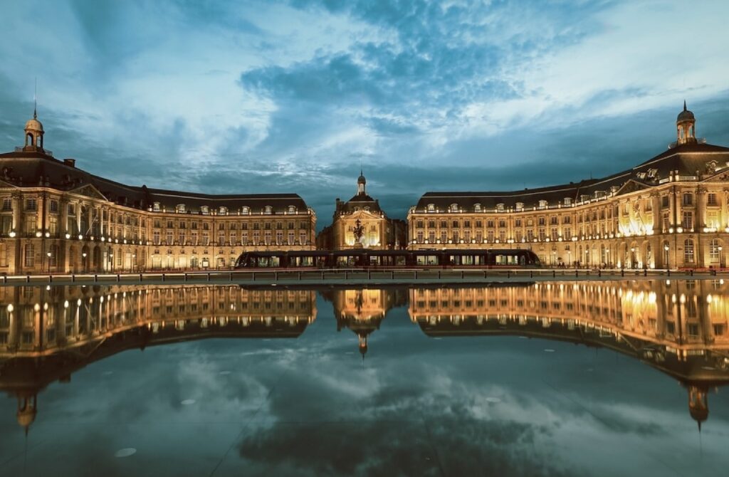 Place de la Bourse in Bordeaux, one of the prettiest cities in France, beautifully illuminated at twilight. The grand building's classical facade reflects symmetrically in the 'Miroir d'eau', creating a stunning visual effect under a dramatic evening sky.
