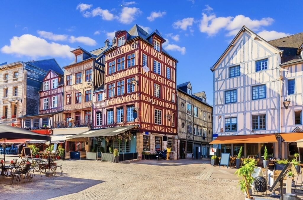 The Old Market Square in Rouen, Normandy, known as one of the prettiest cities in France. The square features vibrant half-timbered houses in red, pink, and white hues, under a clear blue sky. Cafes with outdoor seating add a lively atmosphere to this historic location.