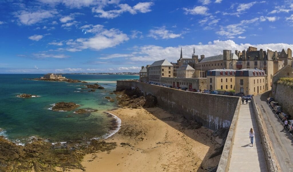 Panoramic view of Saint-Malo, one of the prettiest cities in France, featuring its iconic walled city along the coastline of Brittany. The historic stone walls enclose classic French architecture, with a foreground of a sandy beach and rocky shoreline leading into the azure waters of the English Channel.