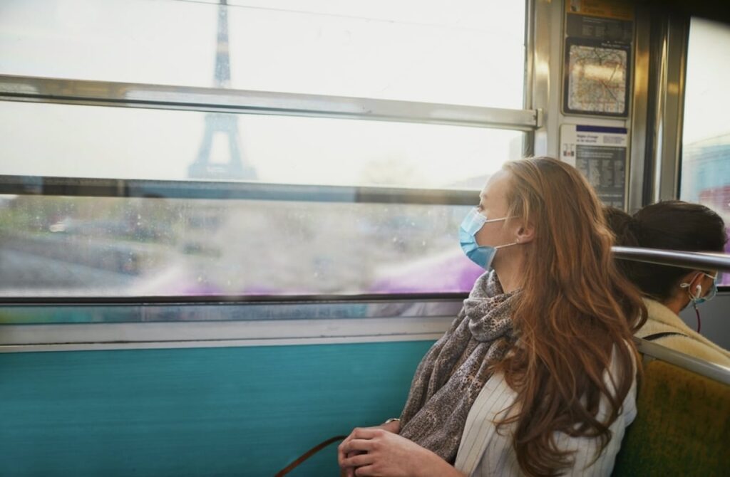 A woman wearing a protective face mask gazes out the window of a Paris metro train, with a blurred view of the Eiffel Tower in the background. She is sitting near another masked passenger, reflecting the precautions taken during the coronavirus pandemic.