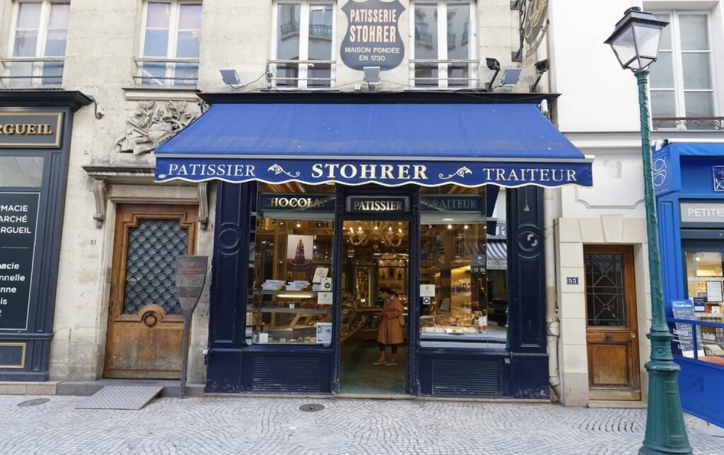 1st arrondissement in paris: The image shows the facade of Stohrer, the oldest cake shop in Paris, founded in 1730, with a blue awning that reads "PATISSIER STOHRER TRAITEUR" and a person browsing inside.
