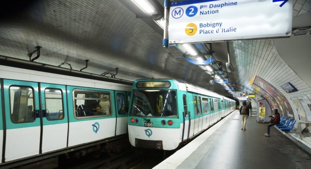 A Paris metro train at a station, with a few passengers waiting on the platform. The station sign indicates connections to metro lines 2 and 5, with destinations including Pte Dauphine, Nation, Bobigny, and Place d'Italie. The station features a tiled arched ceiling and blue seating areas.
