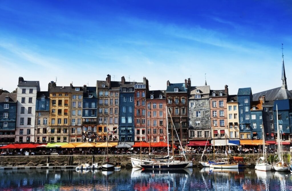 The vibrant waterfront of Honfleur, often celebrated as one of the prettiest cities in France, features a row of colorful, multi-storied buildings. The harbor, filled with boats and surrounded by bustling cafes under bright umbrellas, reflects the lively architectural beauty in its calm waters under a clear blue sky.