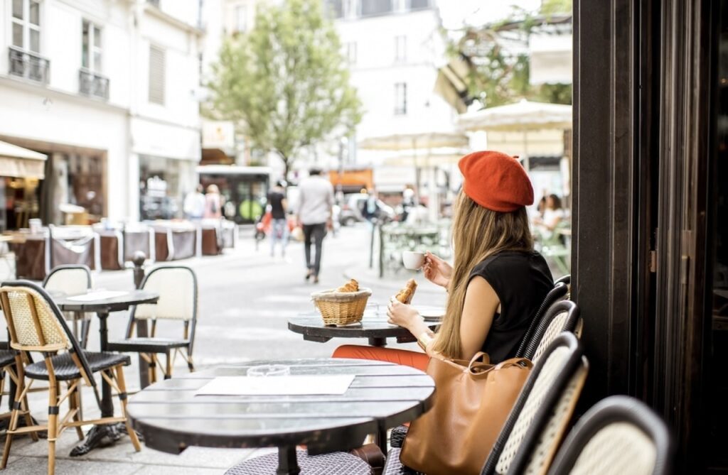 A young woman in a stylish black outfit and a red beret sits at an outdoor café in Paris, enjoying a croissant and coffee. The scene captures the quintessential Parisian street life, bustling with people and lined with other cafés, ideal for Instagram quotes that evoke the timeless charm of Paris.